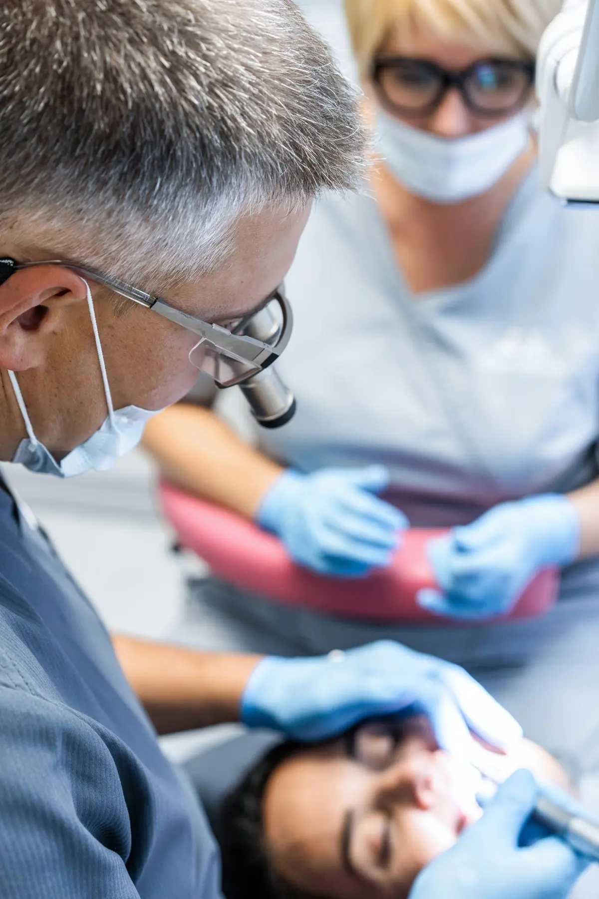 Two masked clinicians leaning over a patient from either side of the chair, one of them guiding the other’s hands