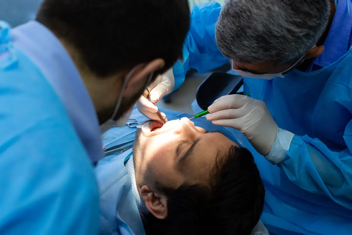 Two operators in blue scrubs and masks bent over a patient, one holding the mirror and the light while the other works