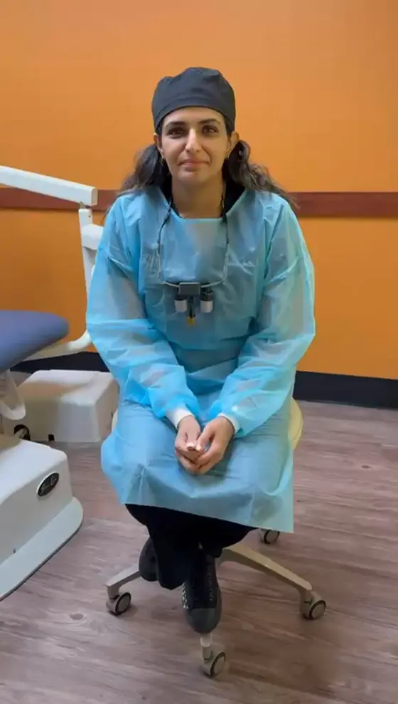 A course participant in a blue gown and surgical cap, loupes hanging from her neck, sitting on a stool in the clinic before recording her testimonial